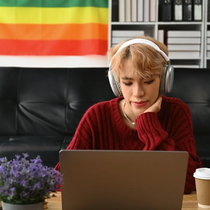 A young gay man wearing headphones works at his laptop with a pride flag on the wall behind him